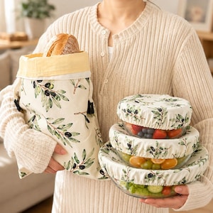 May include: A fabric bread bag with a drawstring closure and a stack of three glass bowls, each covered with a matching elasticated fabric cover. All fabric items feature a cream background with a green olive branch and black olive pattern.