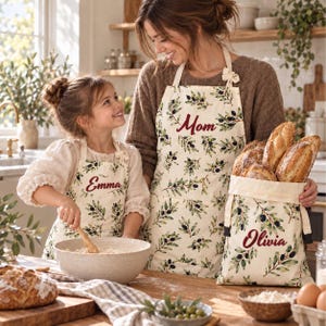 May include: Two matching aprons and a bread bag made of off-white fabric with a green olive branch pattern. The aprons feature the names Emma and Mom in red script, while the bread bag is labeled Olivia in red script and holds several loaves.
