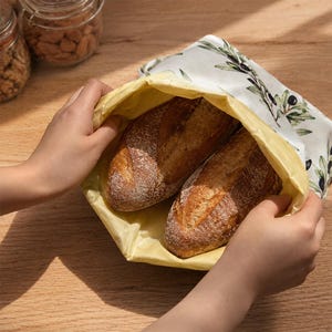 May include: A reusable beeswax food wrap bag featuring a white exterior with a green olive branch pattern and a solid yellow interior, containing two crusty loaves of bread. The flexible fabric wrap is held open to reveal the baked goods inside.
