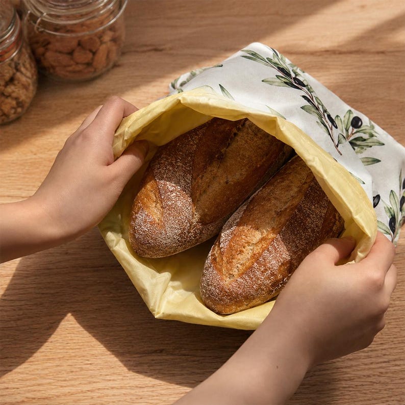 May include: A reusable beeswax food wrap bag featuring a white exterior with a green olive branch pattern and a solid yellow interior, containing two crusty loaves of bread. The flexible fabric wrap is held open to reveal the baked goods inside.