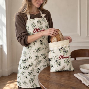 May include: A cream-colored apron and matching bread bag, both featuring a green olive branch pattern and the name Olivia embroidered in red script. The apron has neck straps and the bag has a drawstring closure with a black toggle.