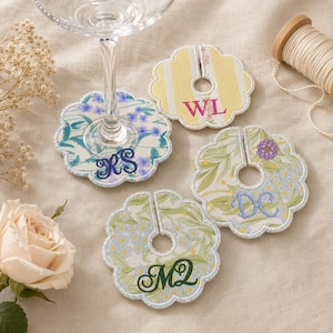 May include: Four scalloped fabric wine glass charms with a central slit. One charm features blue floral patterns with the monogram RS, one has yellow stripes with WL, one has green foliage with DC, and one has green foliage with the monogram MQ.