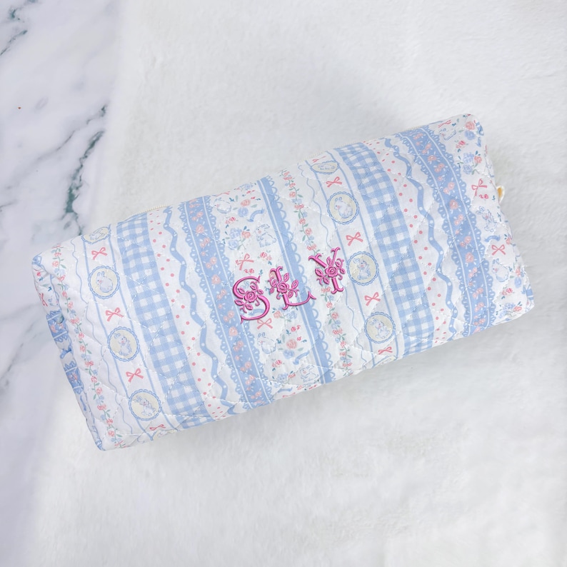 May include: A rectangular, quilted pillow with a blue and white floral and gingham pattern. The pillow features pink embroidered text that reads "SLY". The design includes ribbons and floral accents, creating a soft, feminine aesthetic.