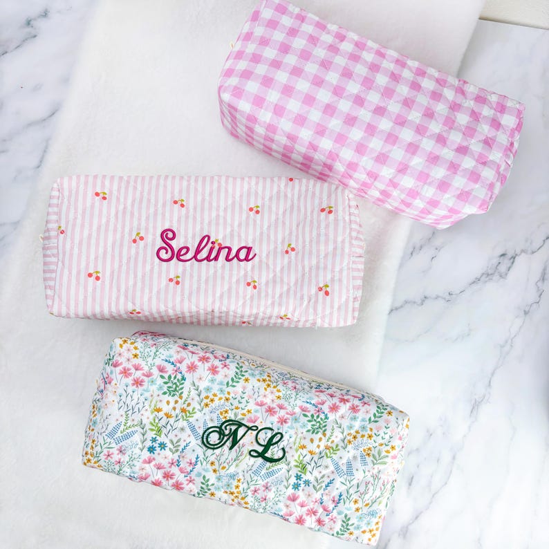 May include: Three quilted cosmetic bags in pink and floral patterns. One bag has pink and white stripes with the name "Selina" embroidered in pink. Another has a pink gingham pattern. The third features a floral design with embroidered initials.