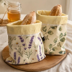 May include: Two fabric storage bags with light yellow interiors and roll-top closures. One bag features a lavender and daisy floral print on a cream background, while the other displays a green eucalyptus leaf pattern. Each bag contains a loaf of bread.