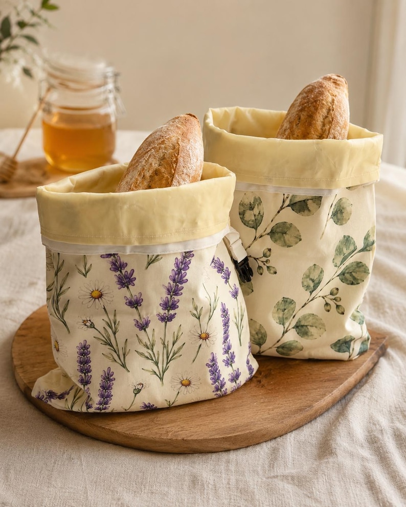 May include: Two fabric storage bags with light yellow interiors and roll-top closures. One bag features a lavender and daisy floral print on a cream background, while the other displays a green eucalyptus leaf pattern. Each bag contains a loaf of bread.