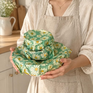 May include: A set of three round bowl covers featuring a green background with a white and yellow daisy floral pattern. The covers are stacked in a tiered arrangement, showing a consistent fabric design across all three sizes of the circular food wraps.