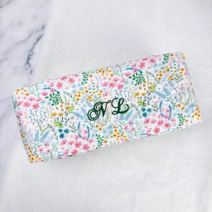 May include: A rectangular, floral-patterned item with a white background. The pattern features pink, yellow, and blue flowers with green leaves. The initials "NL" are embroidered in green on the item.