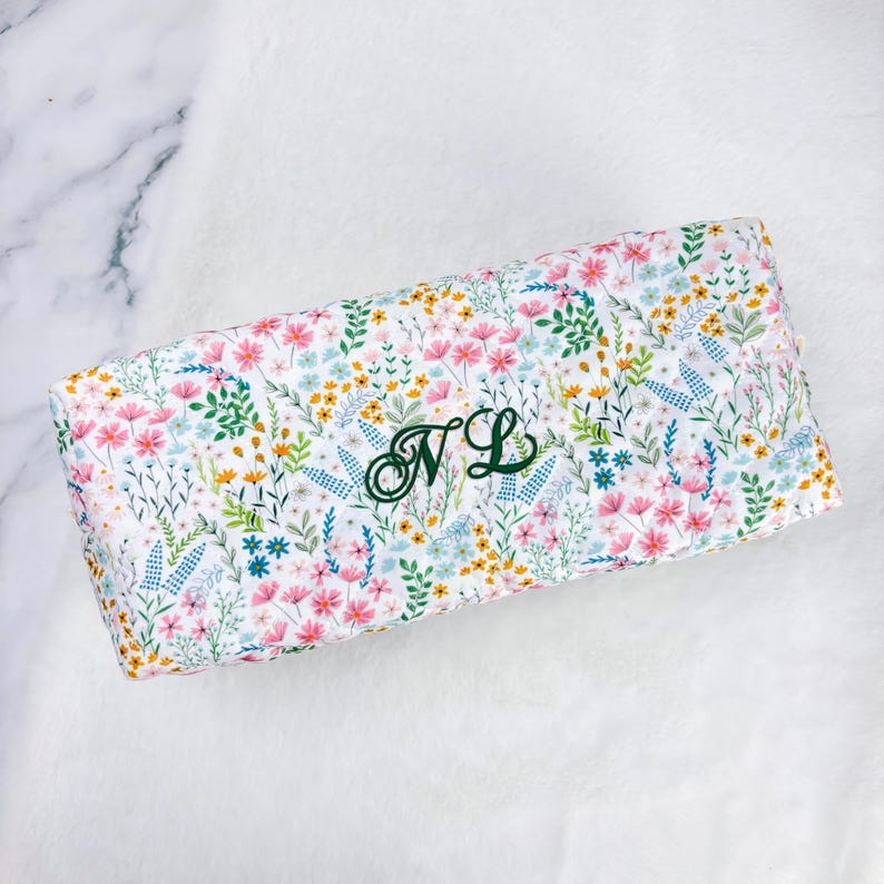 May include: A rectangular, floral-patterned item with a white background. The pattern features pink, yellow, and blue flowers with green leaves. The initials "NL" are embroidered in green on the item.