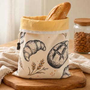 May include: A cream-colored fabric bread bag with a yellow interior lining and a drawstring closure. The exterior features black line-art illustrations of croissants, round loaves of bread, and wheat stalks. A crusty loaf of bread sits inside.