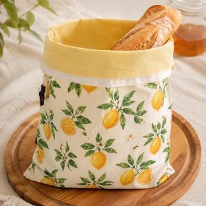 May include: A fabric bread bag featuring a repeating pattern of yellow lemons and green leaves on an off-white background. The bag has a solid yellow interior lining and a black drawstring closure, containing a single crusty baguette loaf.