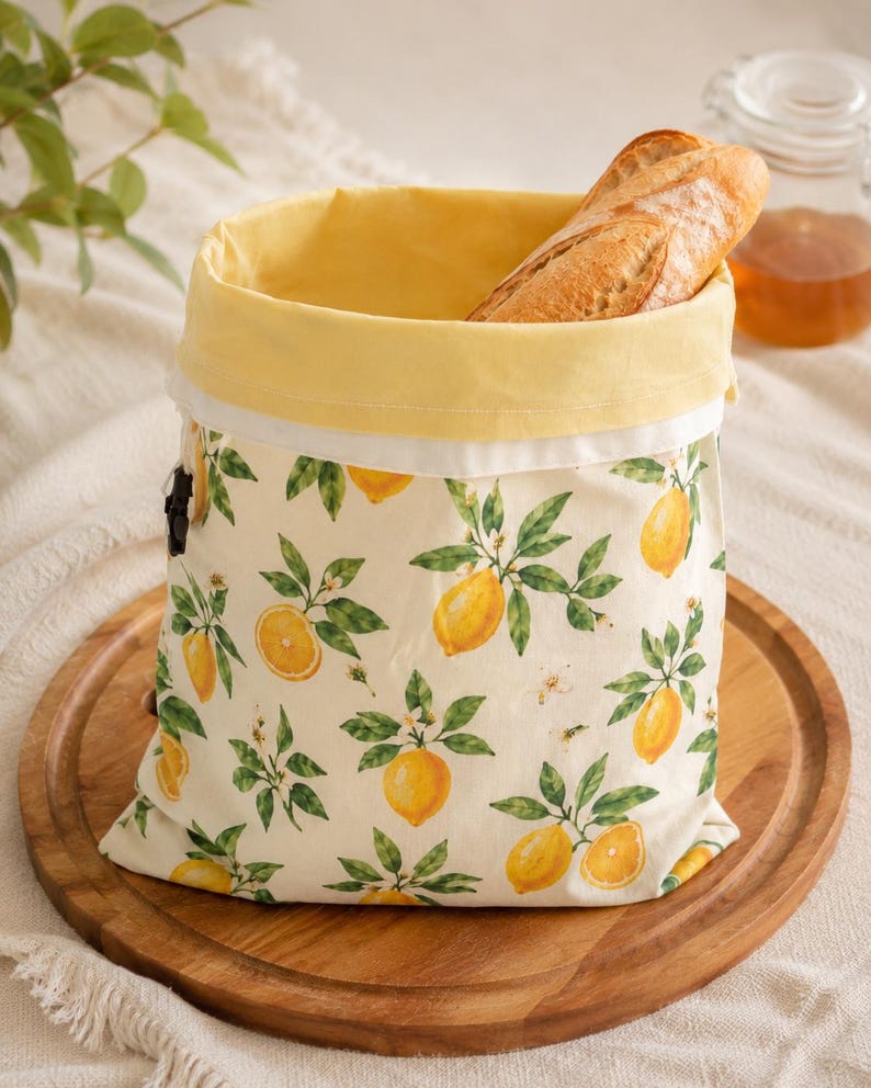 May include: A fabric bread bag featuring a repeating pattern of yellow lemons and green leaves on an off-white background. The bag has a solid yellow interior lining and a black drawstring closure, containing a single crusty baguette loaf.