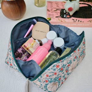 May include: A floral patterned cosmetic bag with a teal zipper and interior, filled with beauty products. The bag features a beige background with green leaves and red flowers. Products include makeup brushes, bottles, and containers. The text "QIJIA BEAUTY" is visible.
