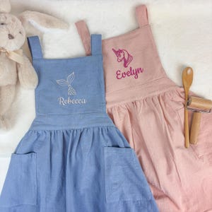 May include: Two aprons, one light blue and one pink, with embroidered designs and names. The blue apron features a mermaid tail and the name "Rebecca." The pink apron has a unicorn and the name "Evelyn." A wooden spoon and rolling pin are next to the pink apron.