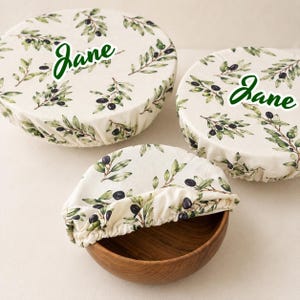 May include: Three round fabric bowl covers with an elastic edge, featuring a cream background with a green olive branch pattern. Two covers are placed flat, and one is fitted over a wooden bowl. The name Jane is printed in green on each cover.