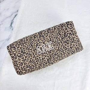 May include: A rectangular, quilted clutch with a leopard print pattern in shades of brown and tan. The clutch features white embroidered initials "SLK" and a gold zipper. The bag is set against a white, textured background.