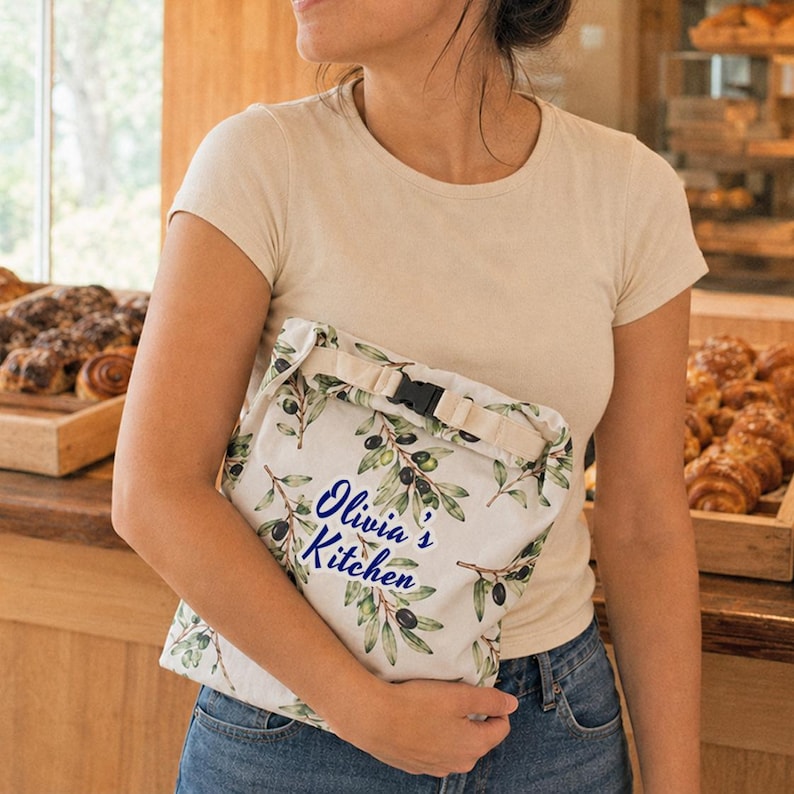 May include: A cream-colored fabric roll-top bag featuring a green olive branch pattern and the blue text "Olivia's Kitchen" printed on the front. The bag includes a beige strap with a black plastic buckle closure for securing the top opening.