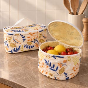 May include: Two fabric storage containers with a cream background and a colorful botanical print featuring blue, orange, and yellow leaves and berries. One container is rectangular with a handle, and the other is round and open, filled with lemons and tomatoes.