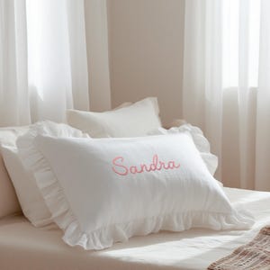 May include: A rectangular white pillow featuring a ruffled border along the edges and the name Sandra embroidered in pink cursive script across the center. The pillow is made of a soft fabric and is positioned on a bed with white linens.