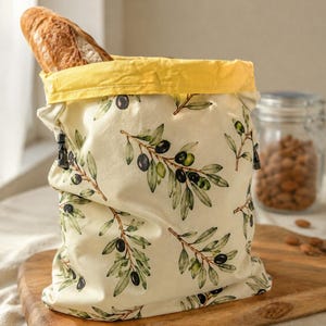 May include: A fabric bread bag with a cream background featuring a green and black olive branch pattern. The bag has a bright yellow top cuff and black drawstring toggles on the sides. A crusty baguette is tucked inside the open top of the bag.