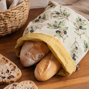 May include: A fabric bread bag with a cream-colored base featuring a repeating pattern of green olive branches and dark olives. The bag has a mustard yellow interior lining and contains two loaves of bread, with additional bread slices nearby.