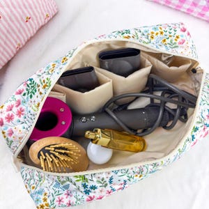 May include: A floral-patterned cosmetic bag filled with beauty tools. The bag contains a hair dryer, brush, perfume, and other accessories. The bag's exterior features a white background with colorful flowers and green leaves.