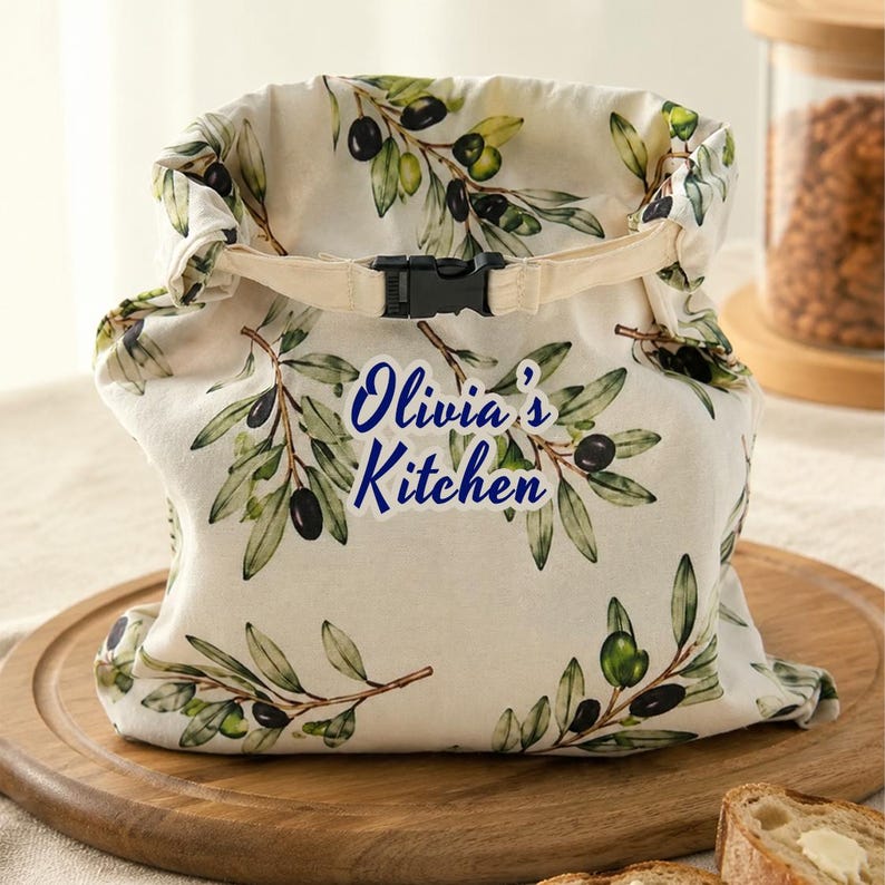 May include: A cream-colored fabric roll-top bag featuring a green olive branch pattern with black olives and leaves. The bag has a black plastic buckle closure at the top and the text "Olivia's Kitchen" printed in blue on the front center.