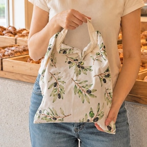 May include: A reusable fabric tote bag featuring an all-over print of green olive branches with dark olives on a cream-colored background. The bag has two integrated fabric handles and a soft, flexible structure, shown held by the top handle.