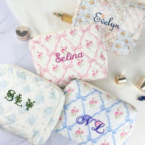 May include: Quilted cosmetic bags in various floral patterns and colors. One bag features the name "Selina" embroidered in pink, while others display initials or names in different colors. The bags have zipper closures.