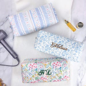 May include: Three rectangular cosmetic bags with floral and patterned designs. One bag is light blue with the name "Melissa" embroidered in brown. Another is white and blue with the initials "NL" embroidered in green. The third bag has a blue and white pattern.