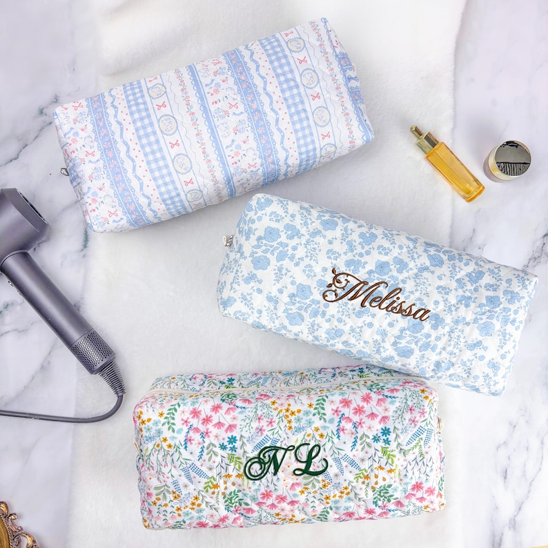 May include: Three rectangular cosmetic bags with floral and patterned designs. One bag is light blue with the name "Melissa" embroidered in brown. Another is white and blue with the initials "NL" embroidered in green. The third bag has a blue and white pattern.