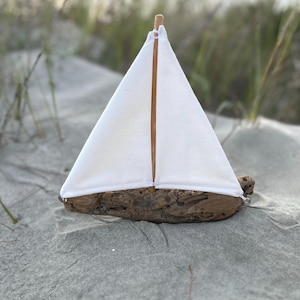 May include: A handcrafted wooden sailboat with a white sail. The boat is made from driftwood and features a triangular sail held up by two wooden dowels. The boat is resting on a sandy surface.