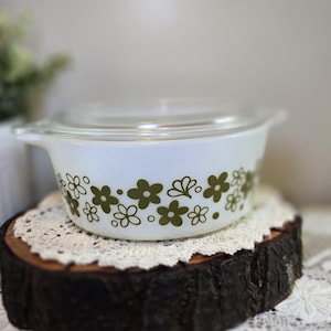May include: White casserole dish with a clear lid, adorned with a green floral pattern. The dish is resting on a wooden slice with a white lace doily. A vintage kitchenware item.