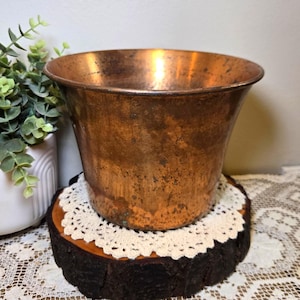 May include: A vintage copper planter with a flared rim, showing a rich patina. The planter sits on a rustic wooden slice with a cream-colored doily. A white ceramic pot with greenery is visible in the background.