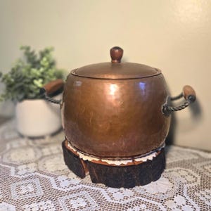 May include: A round, hammered copper pot with a matching lid featuring a dark brown knob. The pot has two twisted metal side handles with brown wooden grips. It rests on a circular wooden base with a dark, textured bark edge and white lace trim.