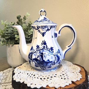 May include: White and blue ceramic teapot with a lid, featuring a detailed blue illustration of a ship and floral patterns. The teapot has a curved spout and handle. It sits on a wooden slice with a white lace doily.