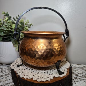 May include: A hammered copper cauldron with a black handle and legs. The cauldron sits on a wooden base with a cream-colored lace doily. The copper pot has a shiny, reflective surface.