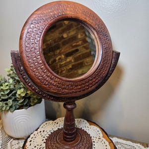 May include: A vintage, round, wooden vanity mirror with a detailed, carved frame and base. The dark brown mirror is mounted on a stand and features a decorative, textured pattern around the frame. The mirror is designed to be adjustable.