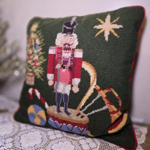 May include: A square dark green needlepoint pillow featuring a central nutcracker figure standing on a drum. The design includes a decorated Christmas tree, a candy cane, a yellow star, and a gold horn. The pillow has a red piped edge.