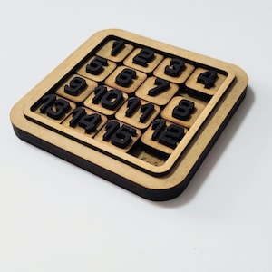 May include: A wooden 15 puzzle game with black numbers on a light brown base. The puzzle is square-shaped, with a frame and sliding tiles. The numbers are arranged from 1 to 15, with one space left open for sliding the tiles.