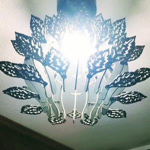 Peut inclure: Un luminaire décoratif de plafond avec un design floral. La lumière est entourée de feuilles découpées au laser en noir et blanc, créant un effet de superposition. Le luminaire est monté sur un plafond blanc.
