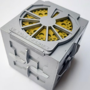 May include: A silver puzzle box with a yellow gear mechanism visible through a circular opening. The box has the words "ULTRA SECRET" engraved on its side. The cube-shaped box has a complex design with interlocking shapes.