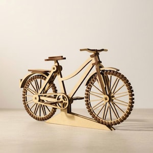 Puede incluir: Modelo de bicicleta de madera con marco marrón claro y detalles en marrón oscuro. El modelo presenta ruedas detalladas, un asiento, manillar y cadena. La bicicleta se exhibe sobre una superficie de color claro, mostrando su intrincado diseño.