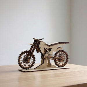 Puede incluir: Modelo de madera de una motocicleta de motocross. El modelo está hecho de madera de color marrón claro y presenta ruedas detalladas, un asiento y un manillar. La motocicleta se exhibe sobre una superficie de madera de color claro.