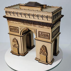 Peut inclure: Maquette en bois détaillée de l'Arc de Triomphe, avec des motifs découpés au laser. La structure beige présente des accents marron foncé et des sculptures en relief détaillées. Le modèle est posé sur une surface blanche.