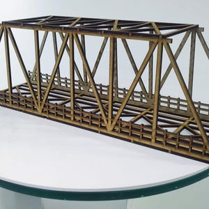 Könnte beinhalten: Ein Holzmodell einer Fachwerkbrücke mit detaillierter Gitterstruktur und Geländern. Die Brücke ist aus hellbraunem Holz gefertigt und wird auf einer weißen Oberfläche präsentiert. Das Modell ist etwa 46 cm lang.