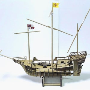 Puede incluir: Maqueta de barco de madera con múltiples mástiles y una cubierta detallada. El barco presenta una bandera amarilla con una cruz y una bandera roja y blanca. El modelo está hecho de madera de color marrón claro y se apoya en una pequeña base.
