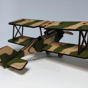 May include: A wooden biplane model with a camouflage pattern of green and tan. The model has two wings, a tail, and a propeller. The biplane is a toy or decorative item.