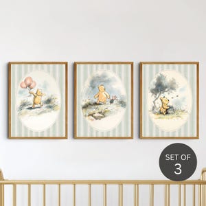 May include: A set of three framed prints, each depicting Winnie the Pooh in different scenes. The illustrations are in a watercolour style, set within oval frames against a striped background. The frames have a warm, wooden finish.