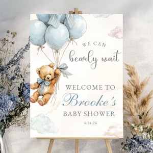 May include: A baby shower welcome sign featuring a teddy bear holding blue balloons. The sign reads "We can bearly wait" and "Welcome to Brooke's Baby Shower". The date 4.14.26 is also displayed.